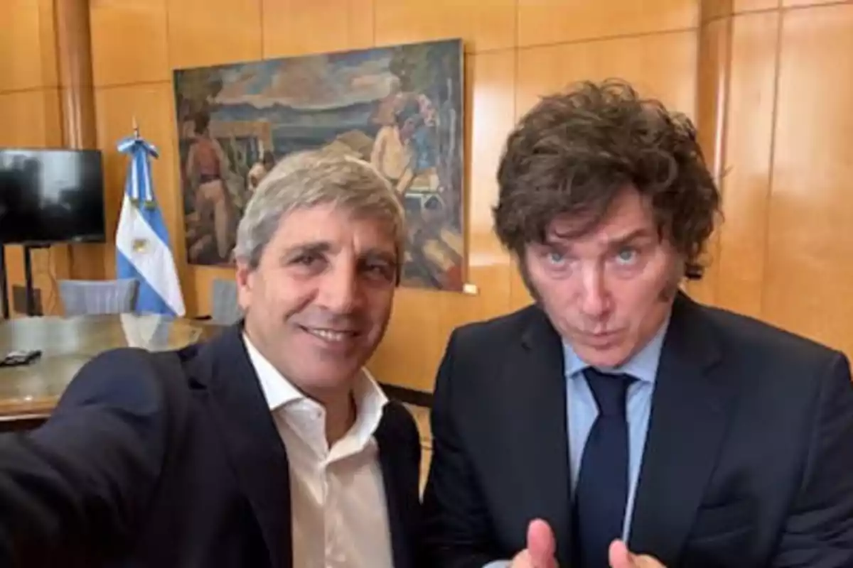 Dos hombres posan para una selfie en una oficina con una bandera argentina y un cuadro al fondo.