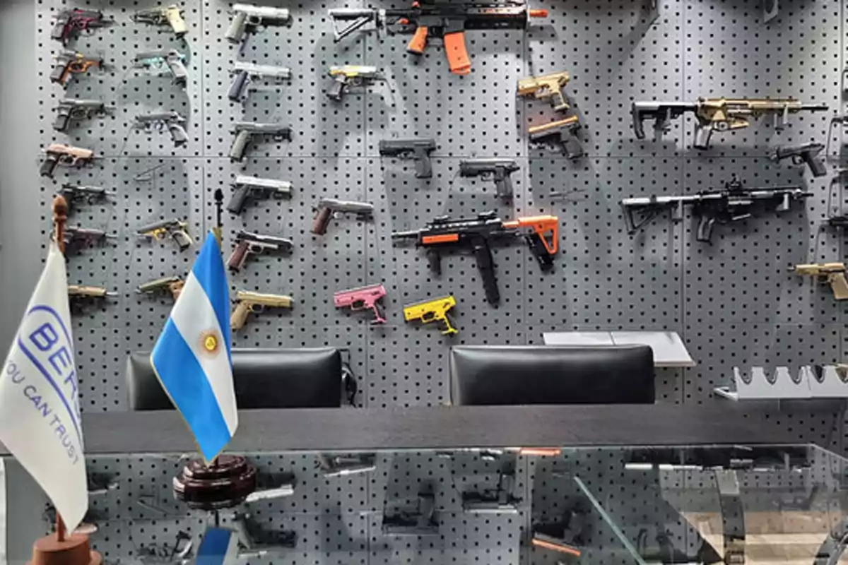 A wall with several firearms displayed on a pegboard with two flags in the foreground.