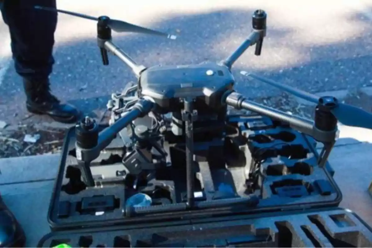 A four-propeller drone is placed on an open transport box on the ground, with a person standing nearby.