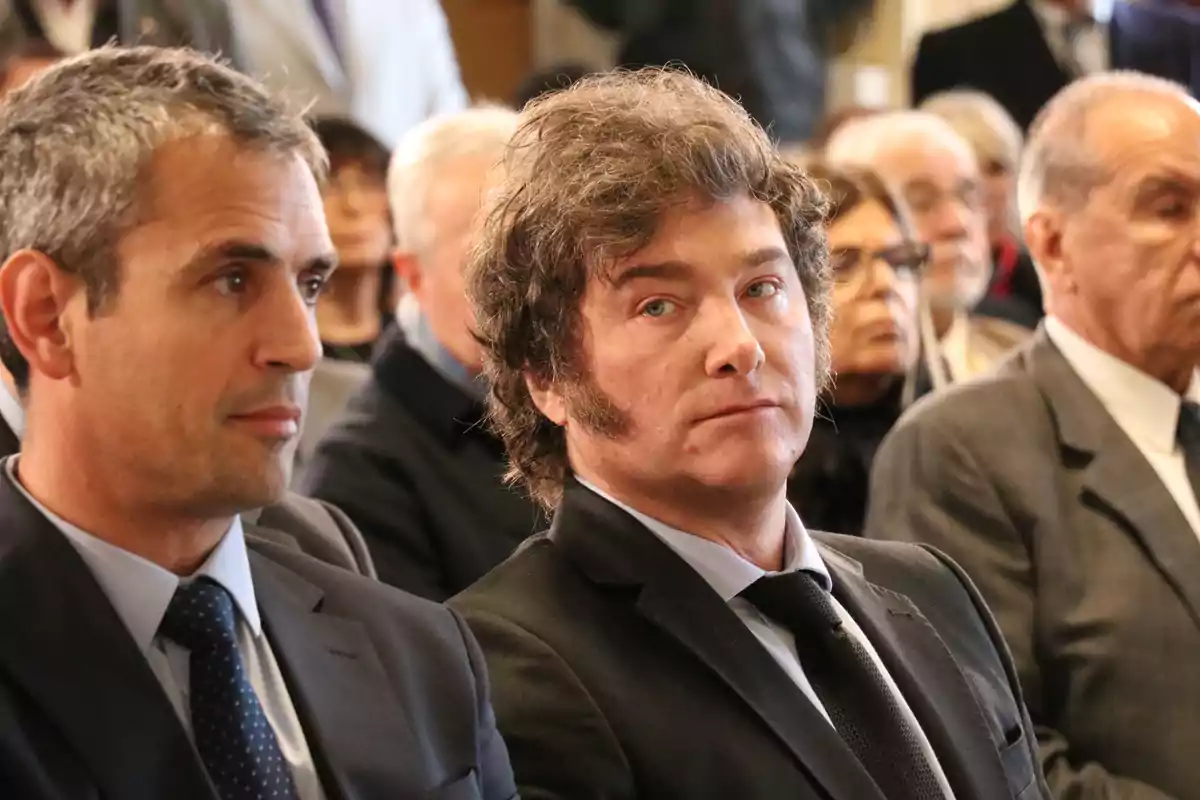 Two men in suits sitting among a group of people at a formal event