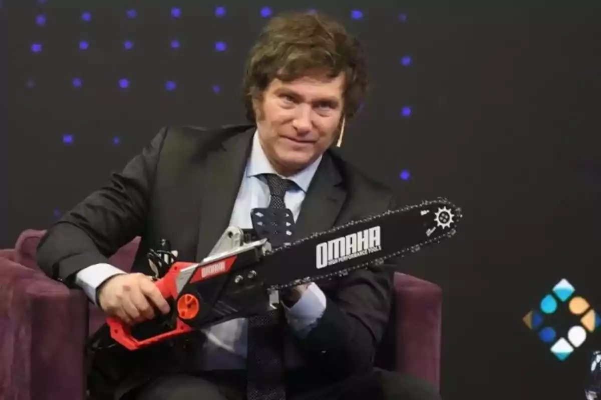 Man in a suit holding a chainsaw sitting in an armchair during a television show