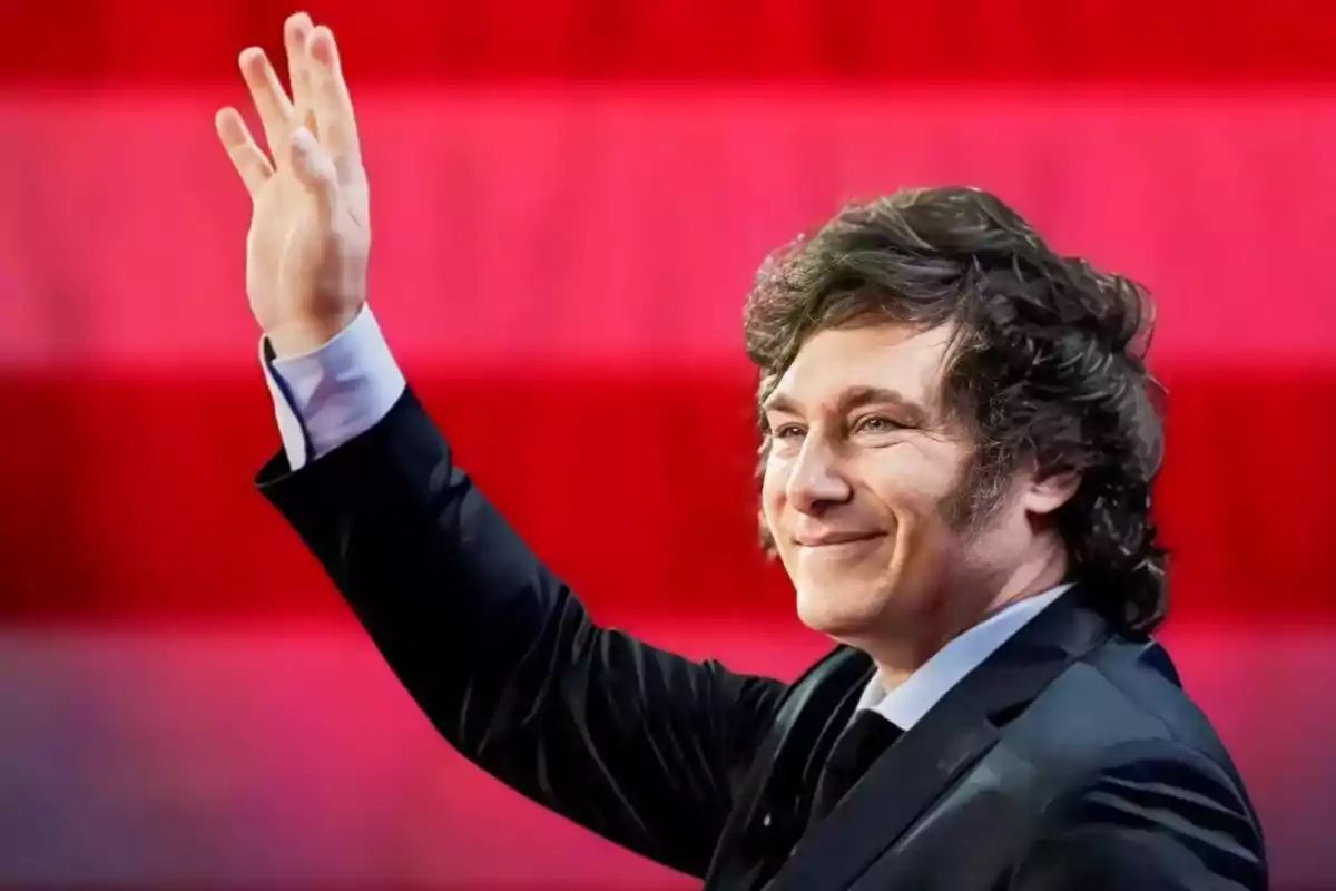 Hombre de cabello oscuro y rizado con traje saludando con la mano levantada frente a un fondo rojo con franjas horizontales