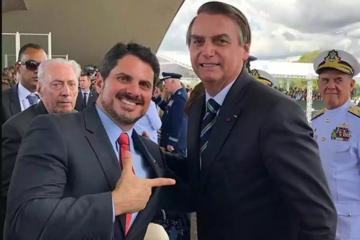 Dos hombres sonrientes posan para una foto en un evento formal, rodeados de otras personas, incluyendo personal militar.