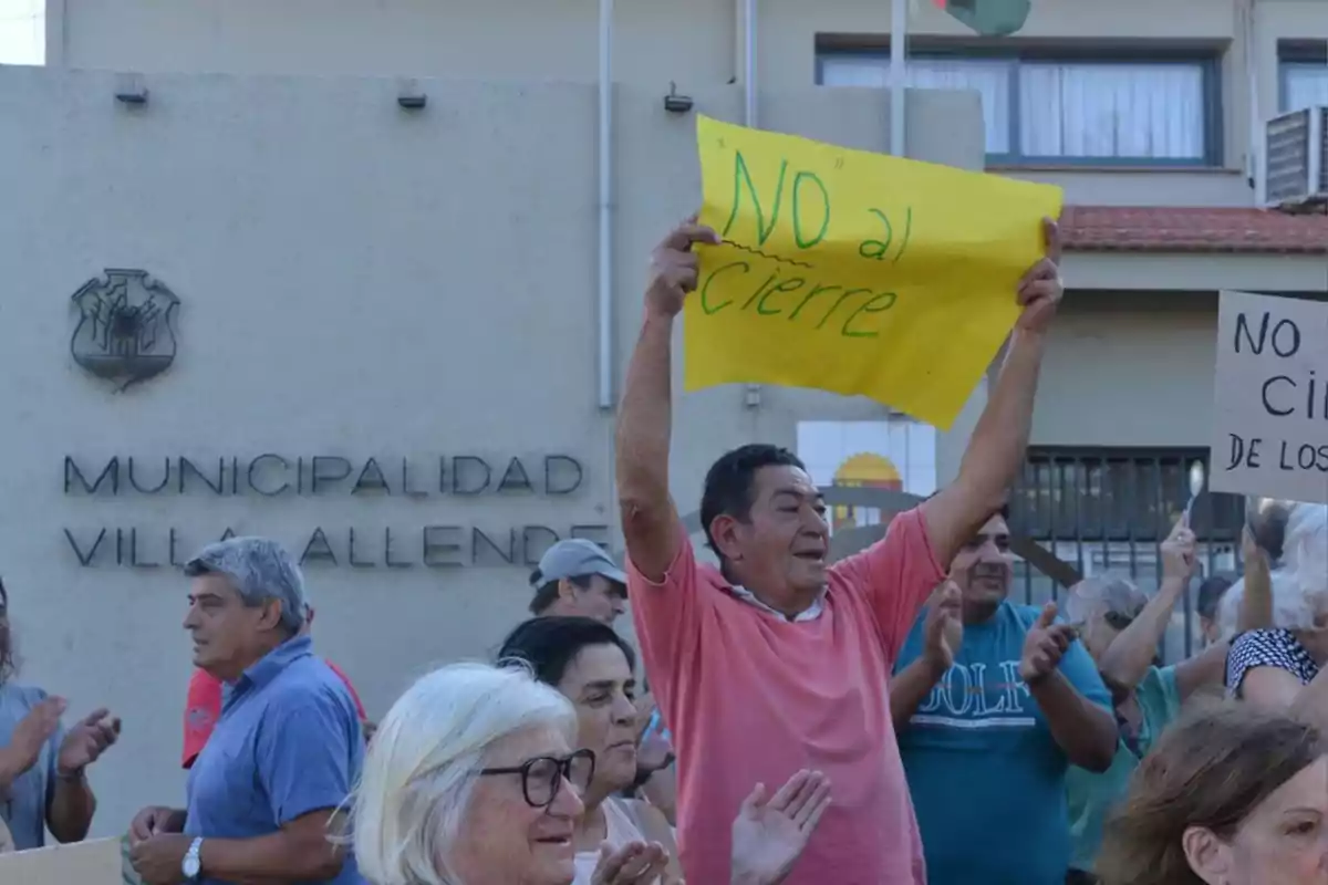 Personas manifestándose frente al edificio de la municipalidad de Villa Allende, un hombre sostiene un cartel amarillo que dice NO al cierre y otras personas aplauden y muestran carteles similares.