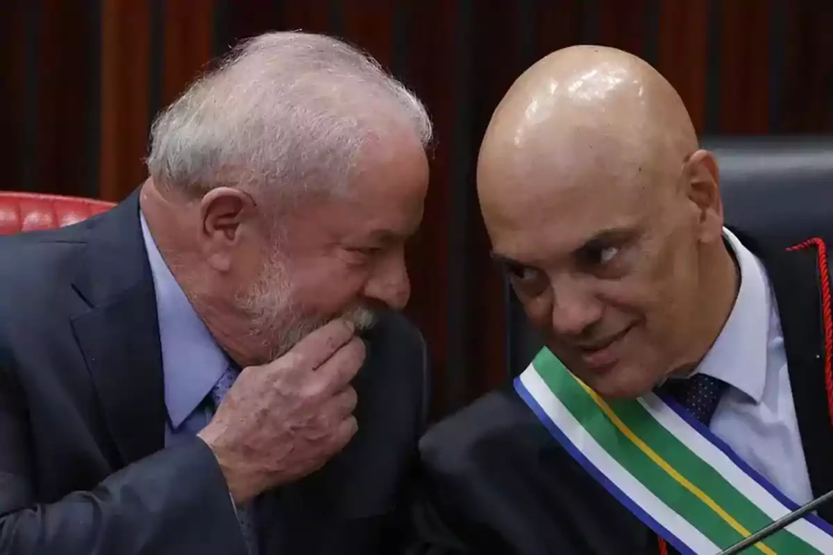 Dos hombres en trajes formales conversan en un entorno oficial, uno de ellos lleva una banda con los colores verde, amarillo y azul.