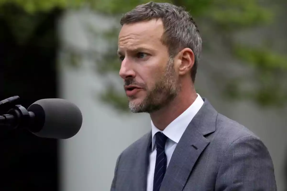 A man with a beard and a gray suit speaks in front of a microphone outdoors.