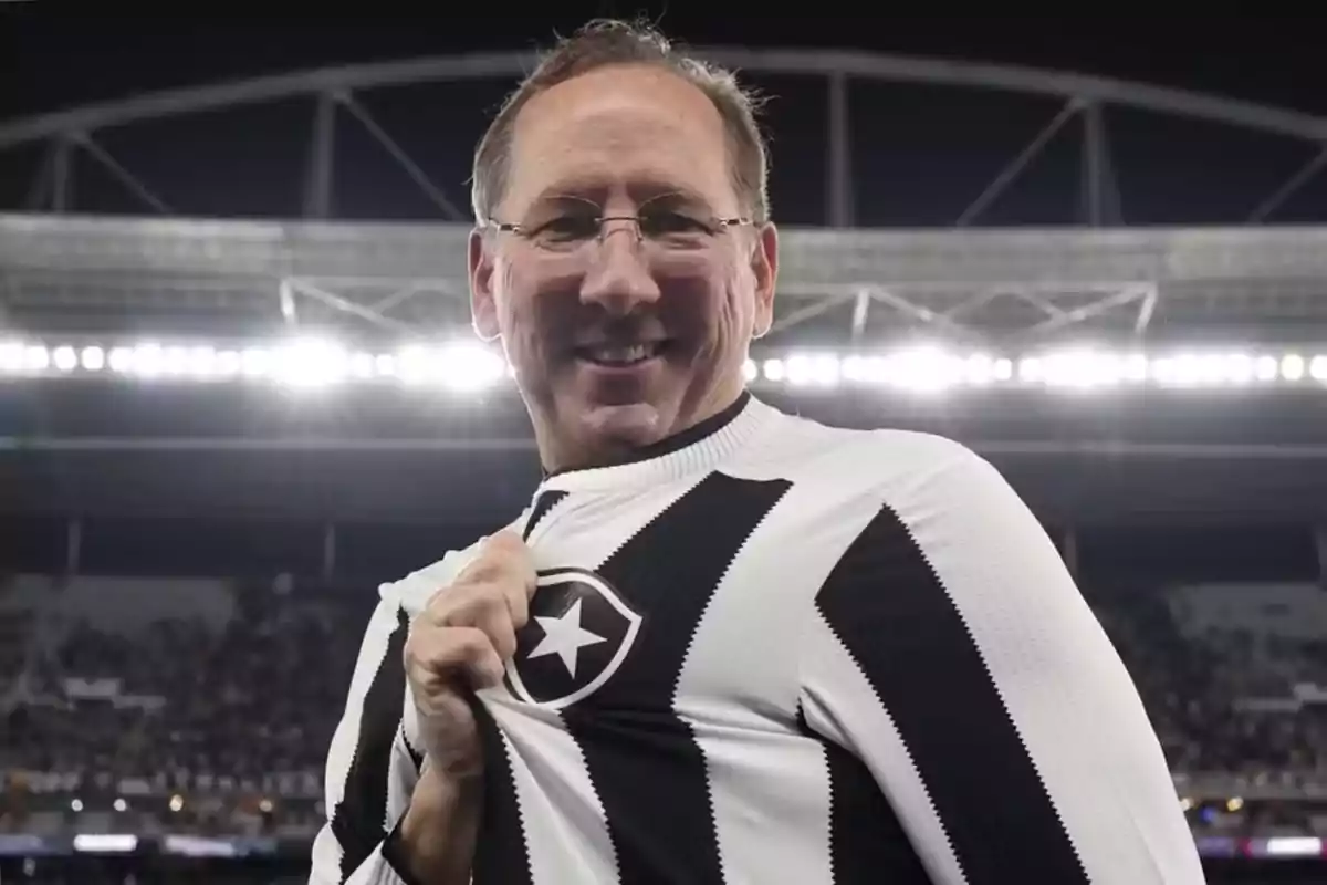 A smiling man with glasses holds a soccer team jersey in a lit-up stadium. A smiling man with glasses holds a soccer team jersey in a lit-up stadium.