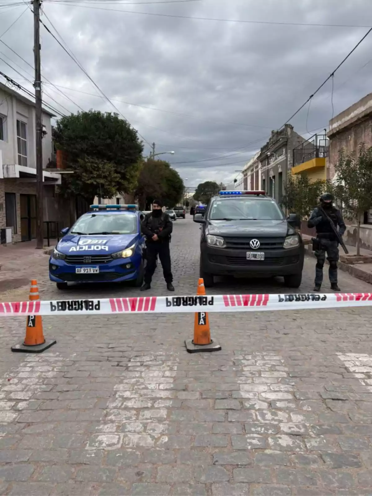 Calle acordonada con cinta de precaución y conos naranjas, dos patrullas policiales y tres agentes de policía custodiando la escena bajo un cielo nublado Calle acordonada con cinta de precaución y conos naranjas, dos patrullas policiales y tres agentes de policía custodiando la escena bajo un cielo nublado