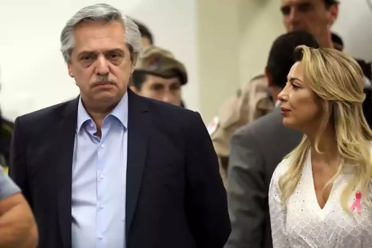 Un hombre de cabello canoso y bigote con expresión seria viste saco oscuro y camisa celeste mientras una mujer rubia con cabello suelto y vestido blanco lo observa en un entorno con varias personas al fondo.