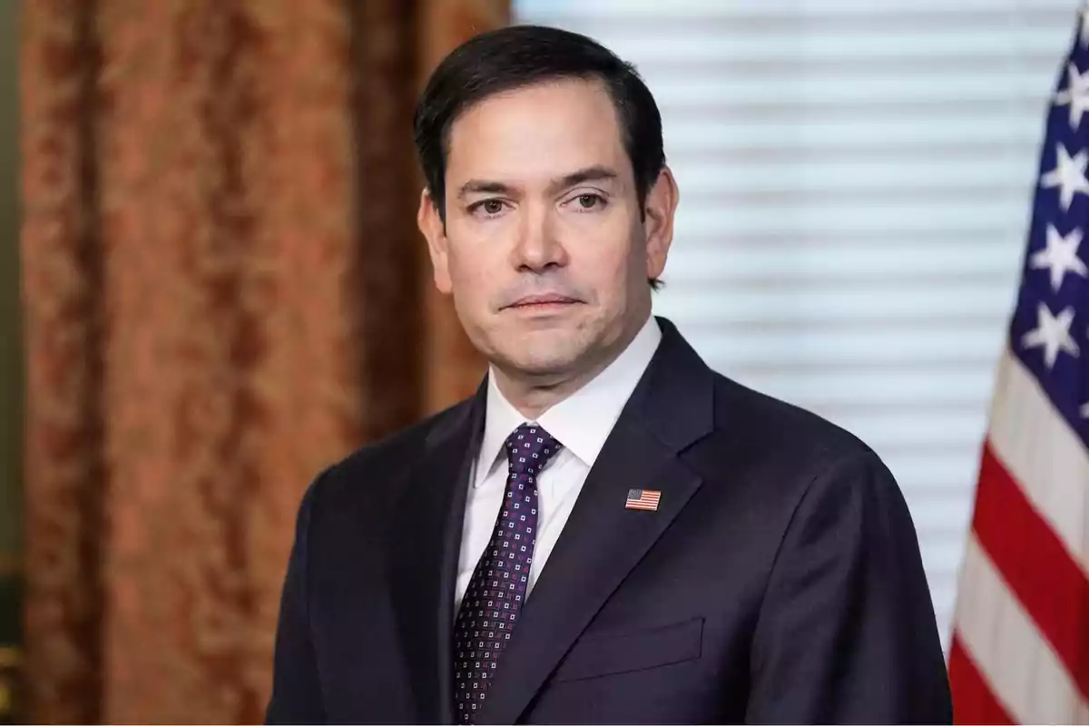 Hombre de cabello oscuro con traje y corbata, lleva un pin de la bandera de Estados Unidos en la solapa y al fondo se ve una bandera estadounidense