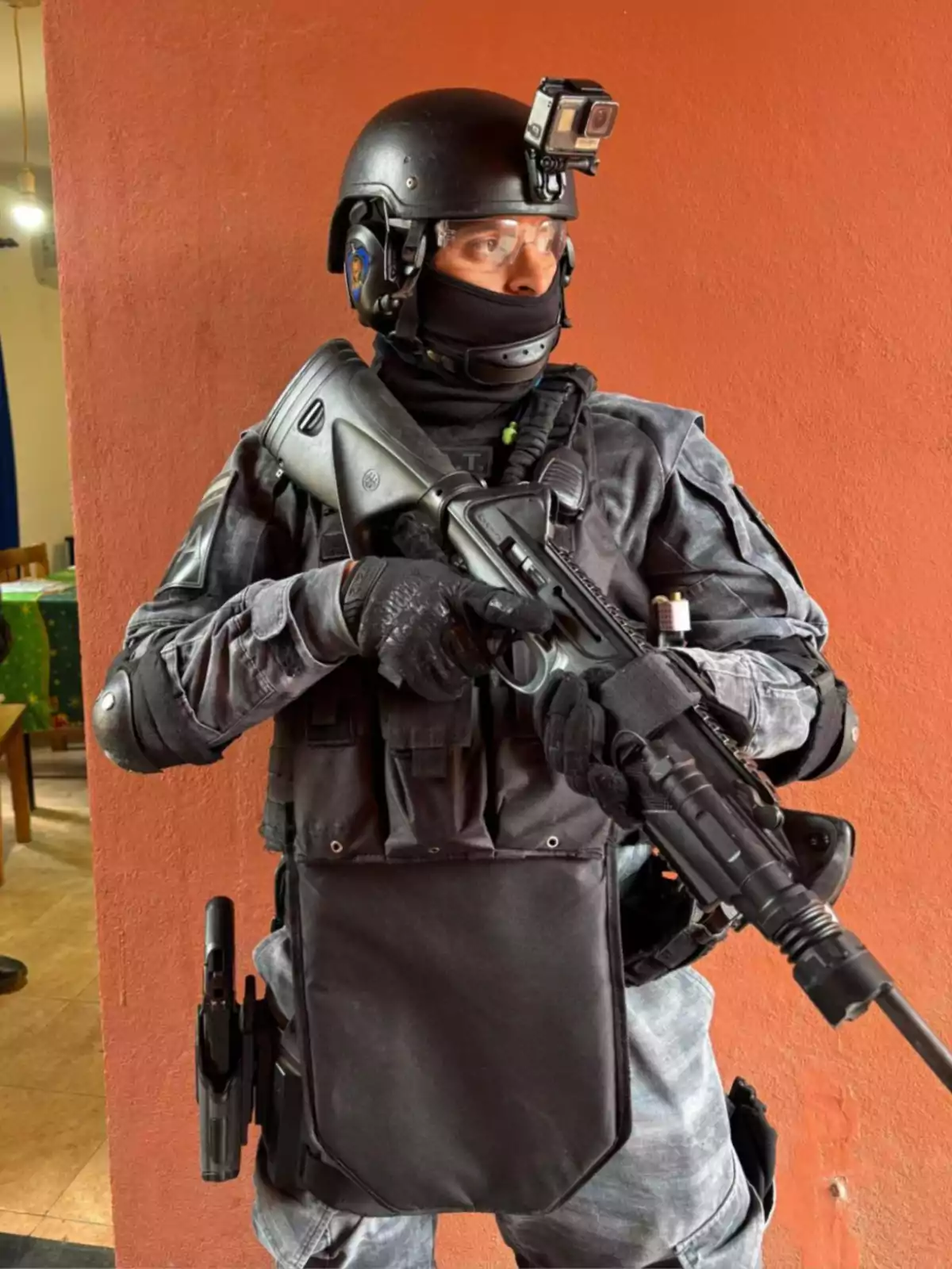 Policía fuertemente armado con uniforme táctico y casco negro sostiene un rifle frente a una pared naranja Policía fuertemente armado con uniforme táctico y casco negro sostiene un rifle frente a una pared naranja