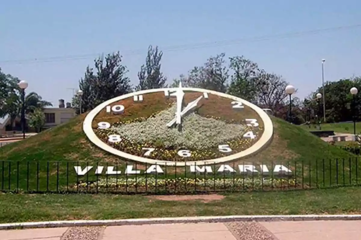 Reloj floral gigante en una colina de césped con la inscripción Villa María en la base y árboles al fondo Reloj floral gigante en una colina de césped con la inscripción Villa María en la base y árboles al fondo