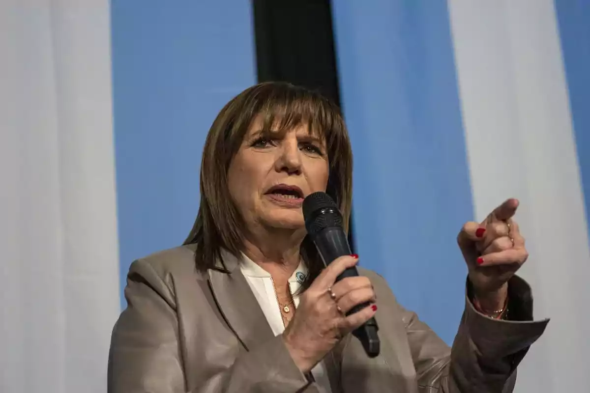 Mujer hablando en público con un micrófono en la mano y fondo de franjas azules y blancas.