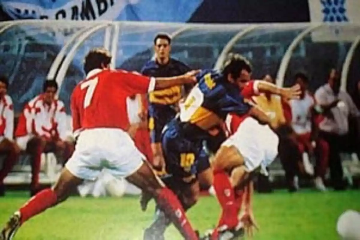 Jugadores de fútbol disputando el balón durante un partido con uno de ellos usando uniforme azul y amarillo y los otros dos con uniforme rojo y blanco Jugadores de fútbol disputando el balón durante un partido con uno de ellos usando uniforme azul y amarillo y los otros dos con uniforme rojo y blanco