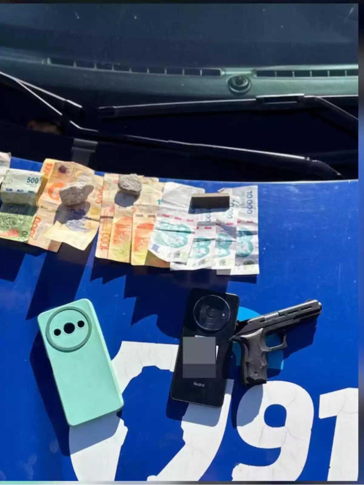 Bills of different denominations, two stones, a small package, two cell phones, and a gun on the hood of a blue vehicle Bills of different denominations, two stones, a small package, two cell phones, and a gun on the hood of a blue vehicle
