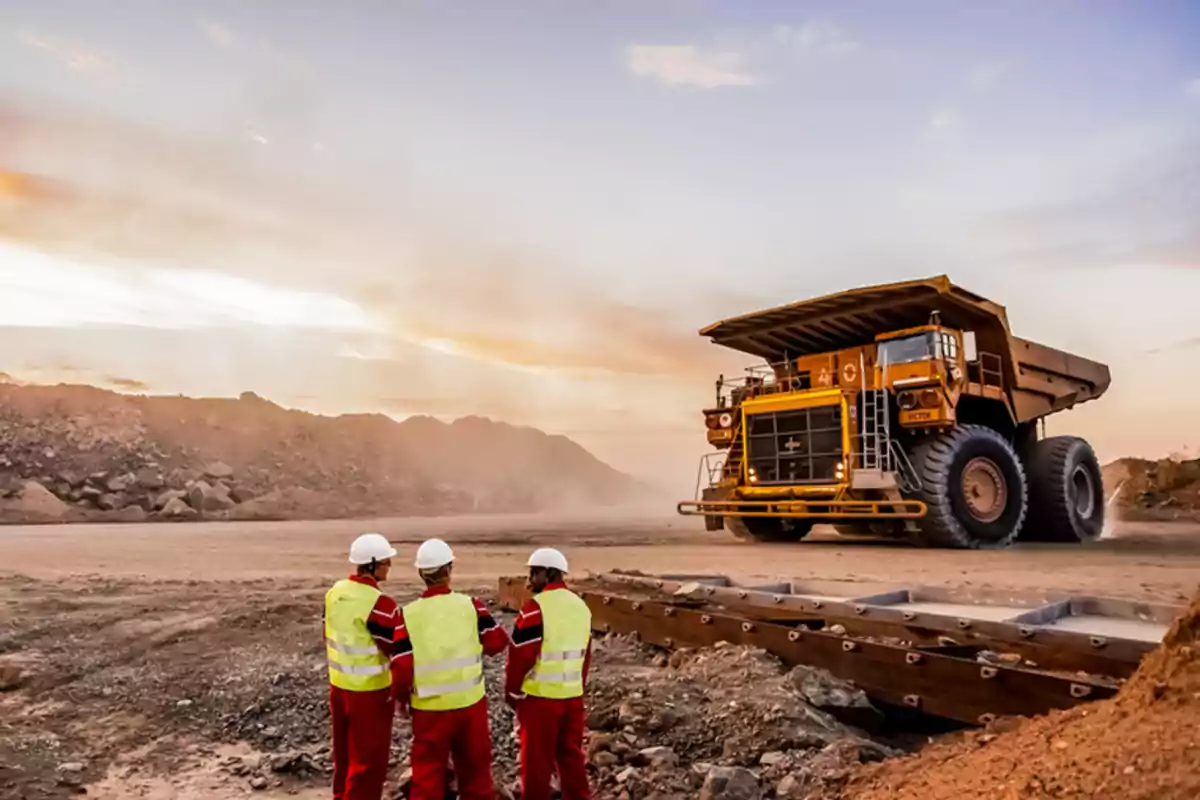Tres trabajadores con cascos y chalecos de seguridad observan un camión de carga grande en un sitio de minería al atardecer. Tres trabajadores con cascos y chalecos de seguridad observan un camión de carga grande en un sitio de minería al atardecer.