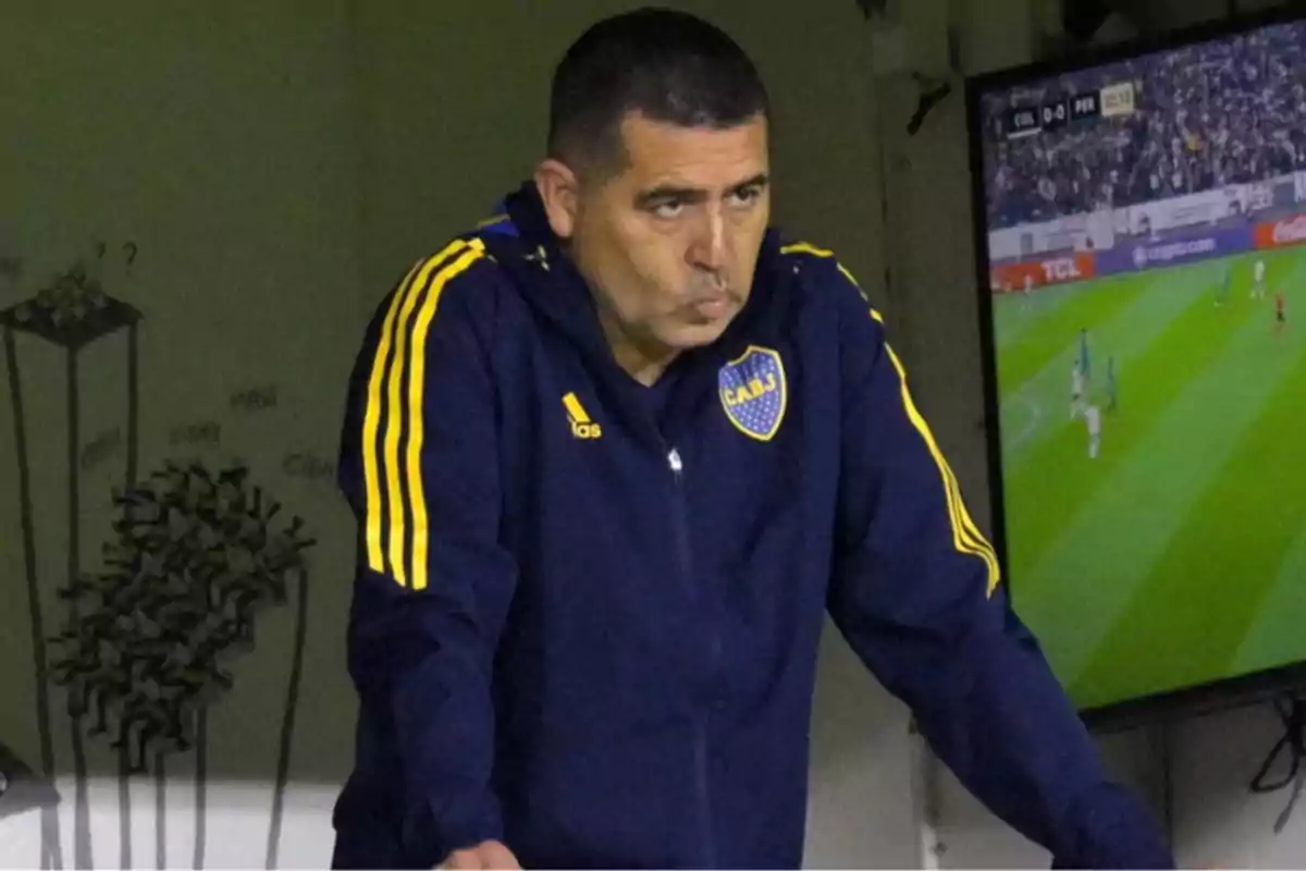 A man in a blue and yellow sports jacket watches a soccer game on TV intently.