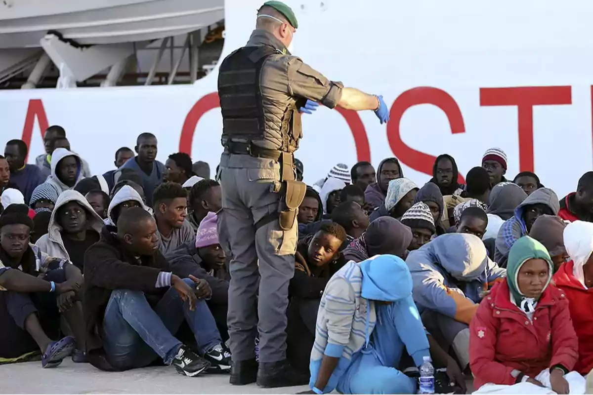 Un grupo de personas sentadas en el suelo junto a un oficial de seguridad en un puerto.