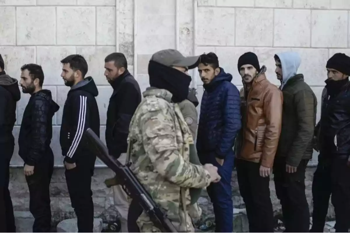 Un grupo de hombres hace fila junto a una pared mientras un soldado armado pasa frente a ellos.