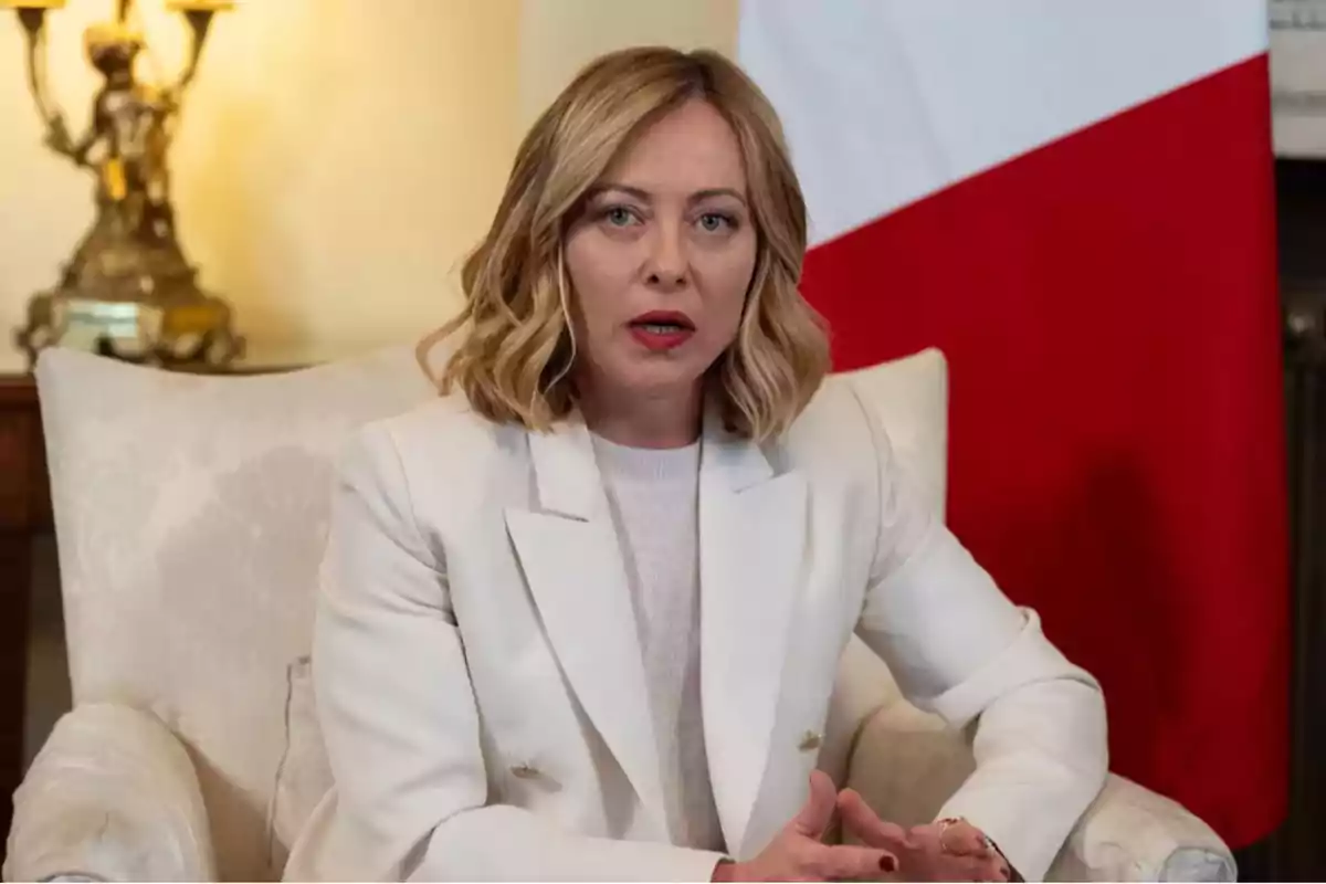 Mujer de cabello rubio y corto sentada en un sillón blanco con una bandera de fondo y expresión seria Mujer de cabello rubio y corto sentada en un sillón blanco con una bandera de fondo y expresión seria