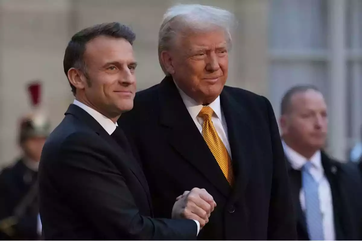 Two men in dark suits shake hands while smiling, with a background of people and a building.