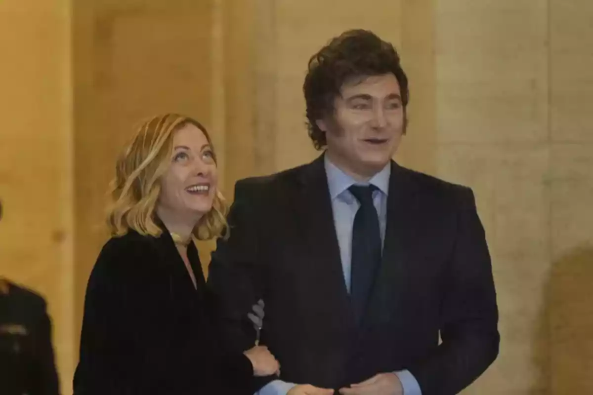 A smiling woman with her arm linked to a man in a dark suit in an indoor setting with light-colored walls