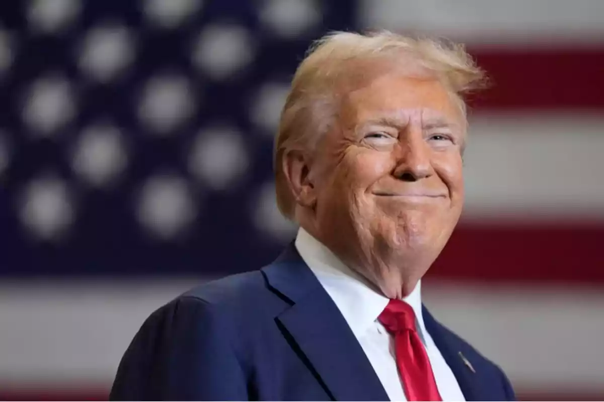 Blond-haired man in a blue suit and red tie smiling in front of a blurred United States flag