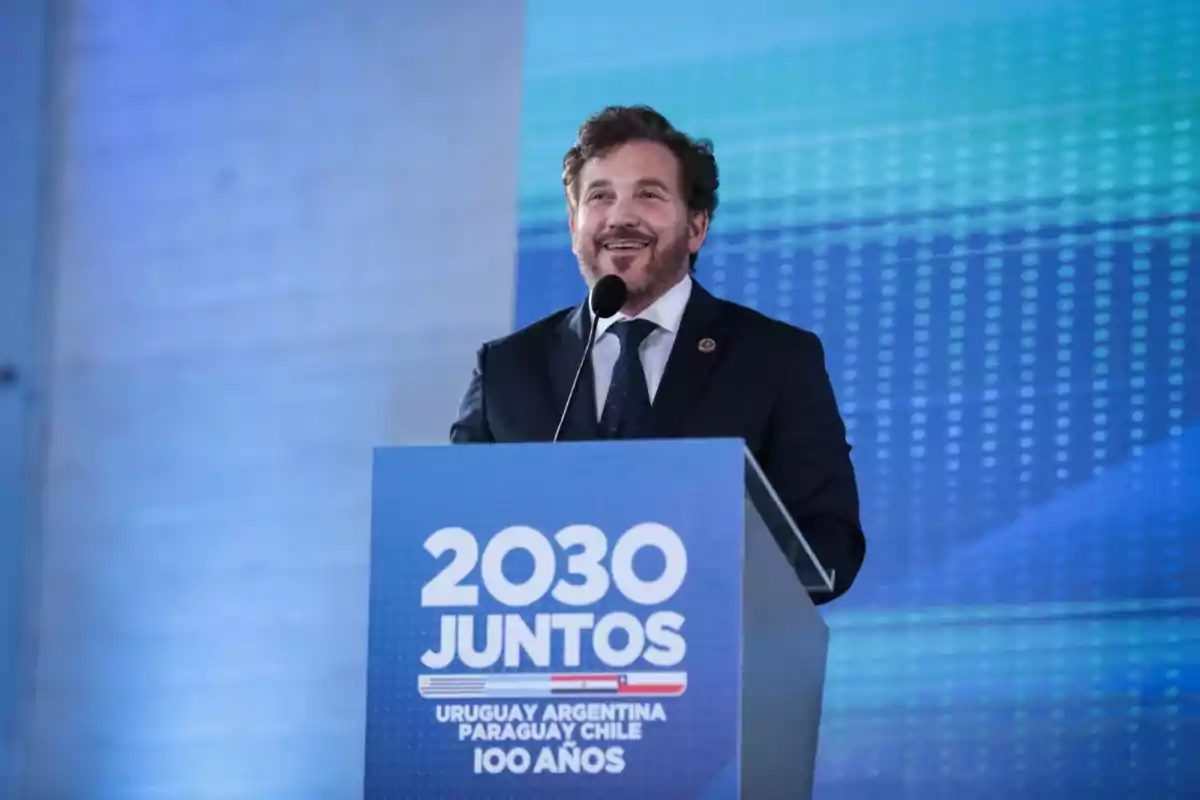Hombre de traje hablando en un podio con un cartel que dice 2030 Juntos Uruguay Argentina Paraguay Chile 100 años sobre un fondo azul Hombre de traje hablando en un podio con un cartel que dice 2030 Juntos Uruguay Argentina Paraguay Chile 100 años sobre un fondo azul