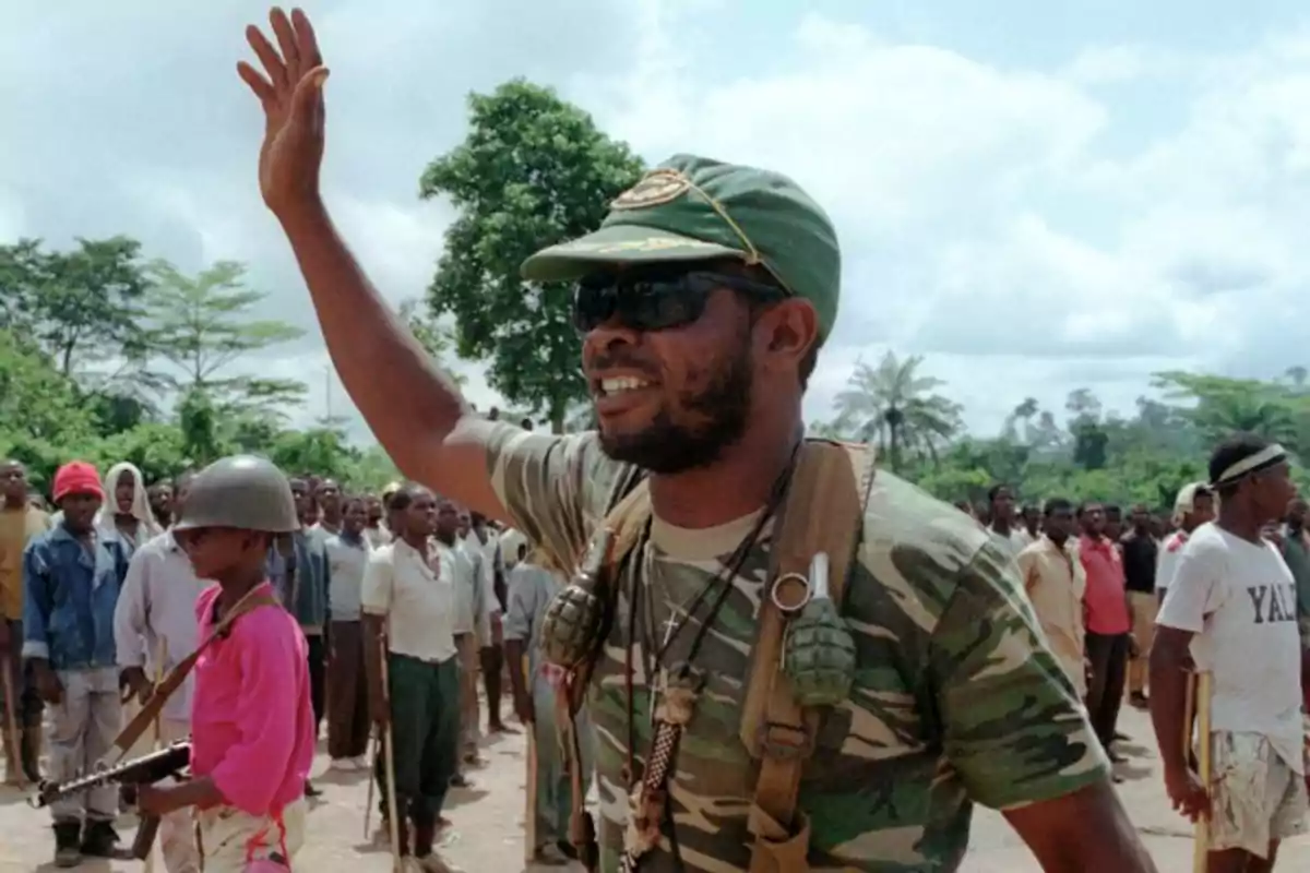 Un hombre con uniforme militar y gafas de sol levanta la mano mientras un grupo de personas, incluyendo un niño con casco y arma, se encuentra detrás de él en un entorno al aire libre con árboles. Un hombre con uniforme militar y gafas de sol levanta la mano mientras un grupo de personas, incluyendo un niño con casco y arma, se encuentra detrás de él en un entorno al aire libre con árboles.