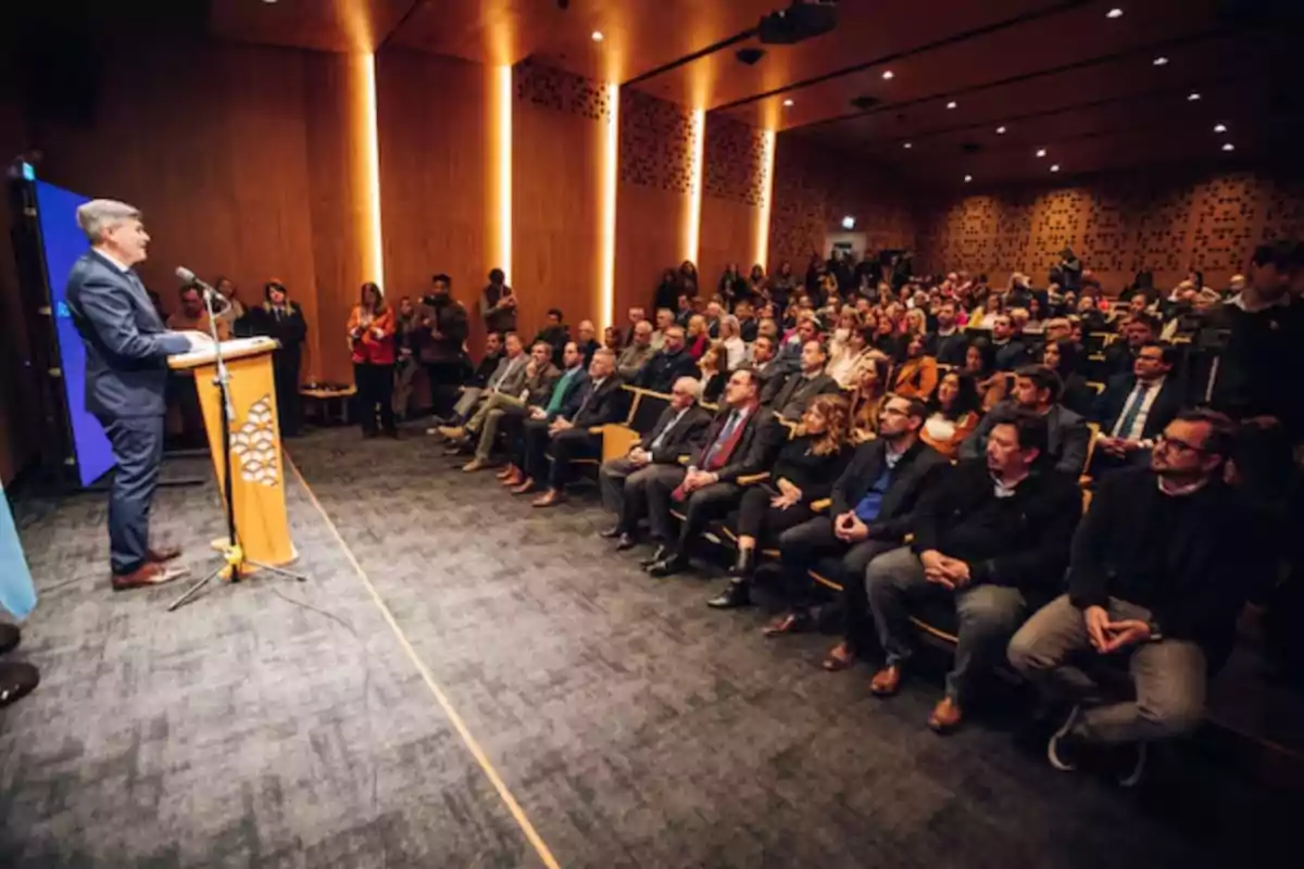 A man in a suit speaks at a podium in front of a large audience in a modern auditorium with wooden walls and warm lighting. A man in a suit speaks at a podium in front of a large audience in a modern auditorium with wooden walls and warm lighting.