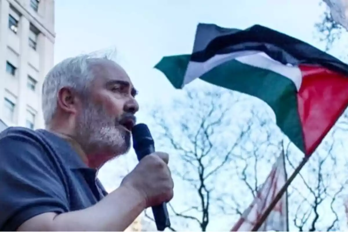 Hombre hablando por un micrófono en una manifestación con una bandera en el fondo.