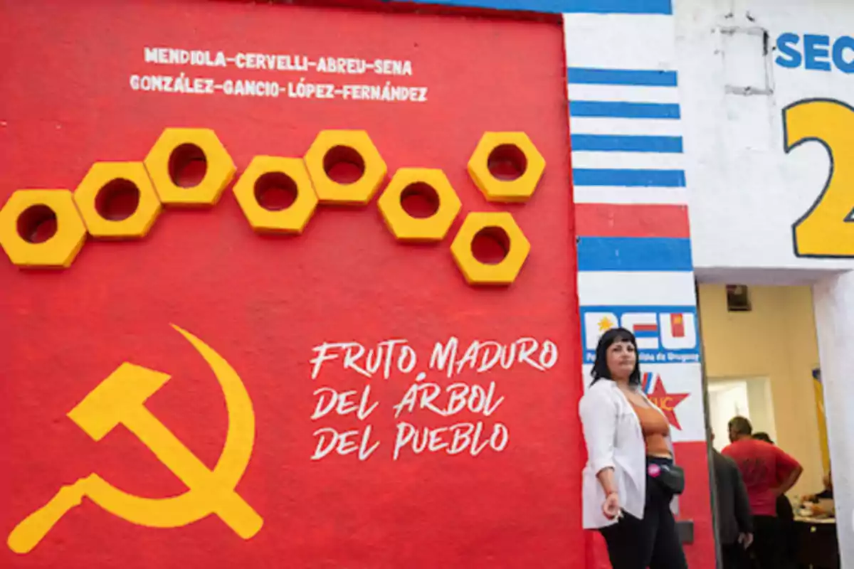 Red mural with yellow nuts, the sickle and hammer, a phrase in Spanish, and a woman standing next to the entrance of a building Red mural with yellow nuts, the sickle and hammer, a phrase in Spanish, and a woman standing next to the entrance of a building