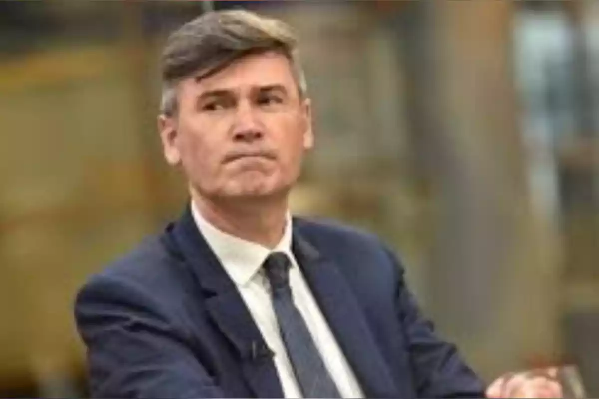 Short-haired, gray-haired man wearing a blue suit and tie, with a serious expression in a blurred indoor setting Short-haired, gray-haired man wearing a blue suit and tie, with a serious expression in a blurred indoor setting