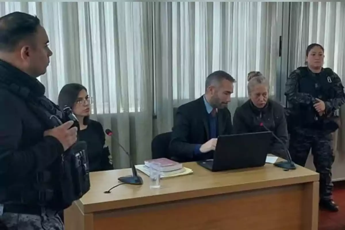 Cinco personas en una sala de audiencias, dos de ellas con uniforme policial, una mujer mayor y un hombre con computadora portátil sentados en una mesa, y una mujer joven con gafas sentada al fondo. Cinco personas en una sala de audiencias, dos de ellas con uniforme policial, una mujer mayor y un hombre con computadora portátil sentados en una mesa, y una mujer joven con gafas sentada al fondo.