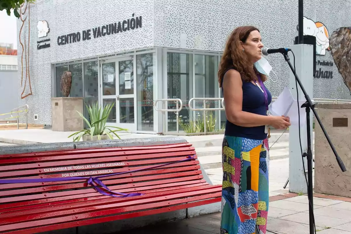 Woman speaking in front of a microphone next to a red bench with the phrase in memory of all the women murdered by those who claimed to love them, in front of a vaccination center.