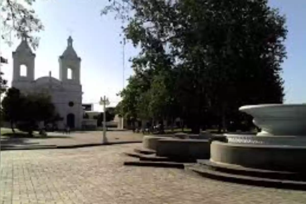 Plaza con una fuente grande en primer plano, árboles y una iglesia blanca al fondo en un día soleado Plaza con una fuente grande en primer plano, árboles y una iglesia blanca al fondo en un día soleado