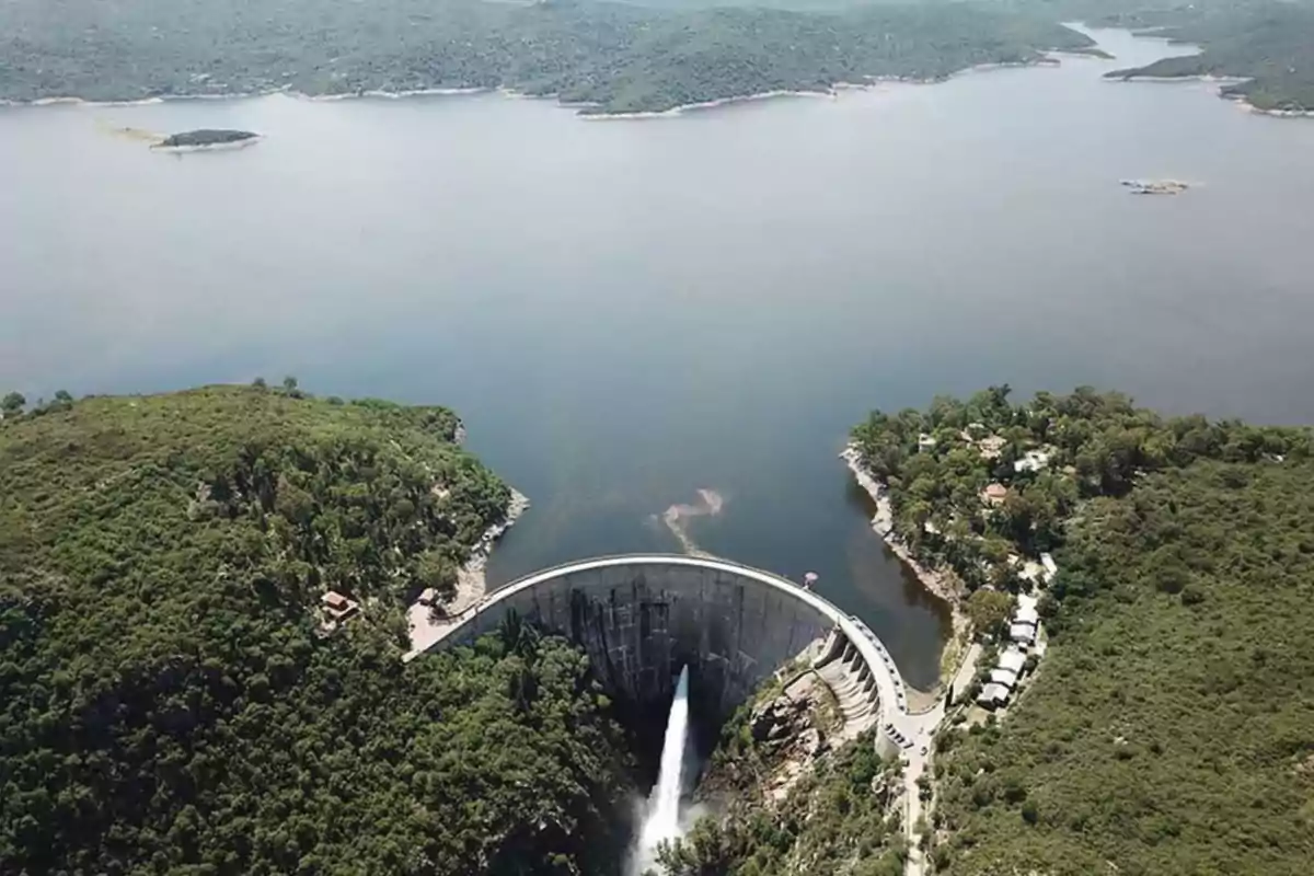 Vista aérea de una represa rodeada de vegetación con agua saliendo por el vertedero y un lago extenso al fondo Vista aérea de una represa rodeada de vegetación con agua saliendo por el vertedero y un lago extenso al fondo
