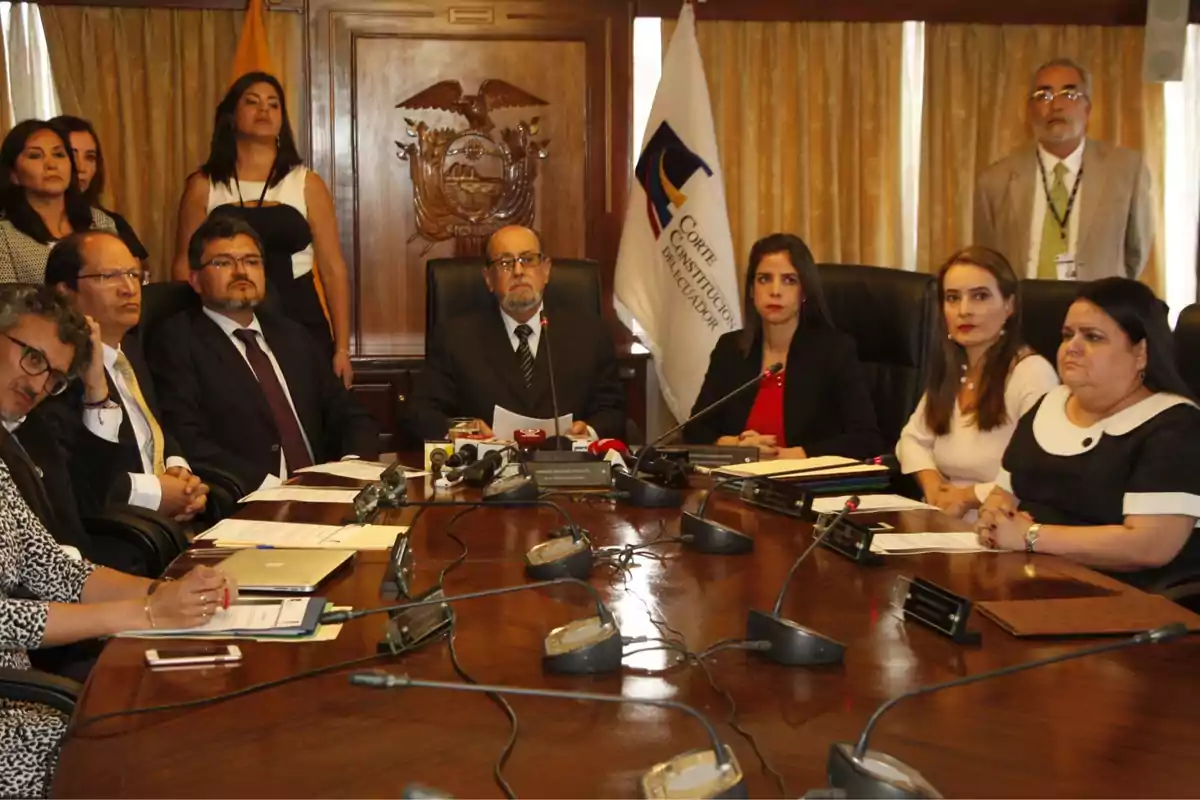 Un grupo de personas en una reunión formal alrededor de una mesa con micrófonos y documentos, en una sala con un emblema y una bandera en el fondo.