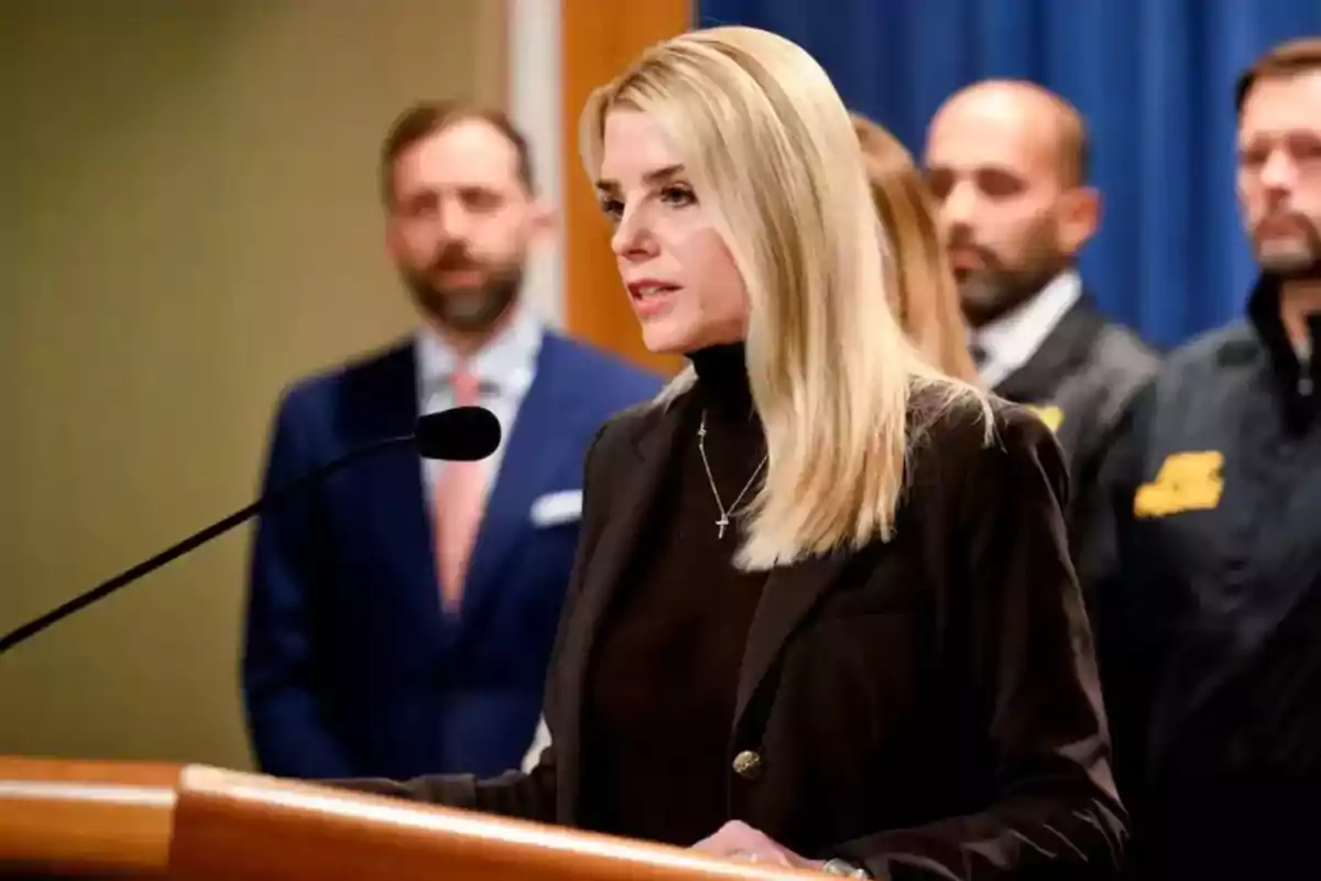 Una mujer rubia hablando en un podio con un micrófono mientras varias personas están de pie en el fondo.