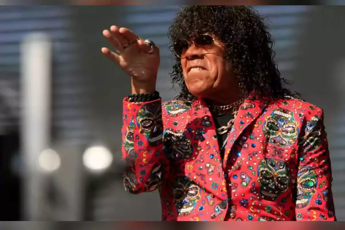 Hombre con cabello rizado y gafas oscuras usando un saco rojo decorado con calaveras coloridas y haciendo un gesto con la mano