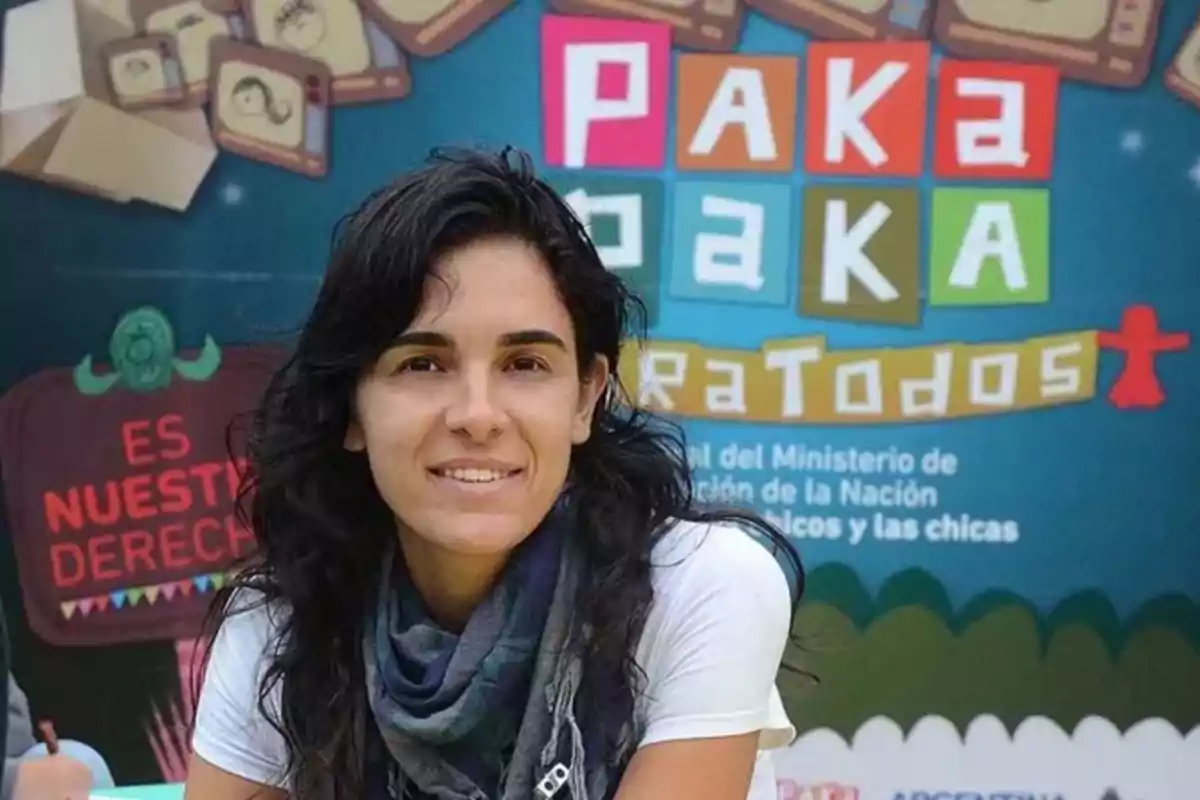 Una persona sonriente con cabello oscuro y una bufanda está frente a un cartel colorido que dice 