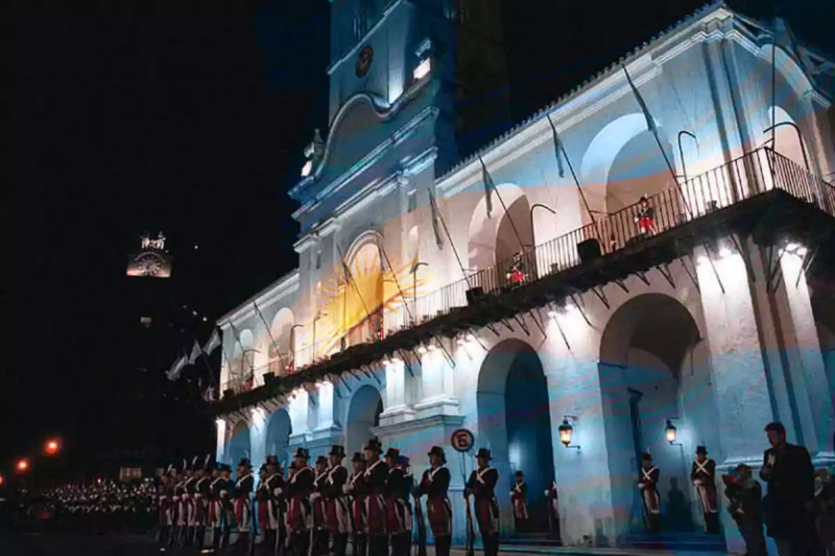 A historic building illuminated at night with a flag projected on its facade and a group of people dressed in traditional uniforms in formation in front of it. A historic building illuminated at night with a flag projected on its facade and a group of people dressed in traditional uniforms in formation in front of it.
