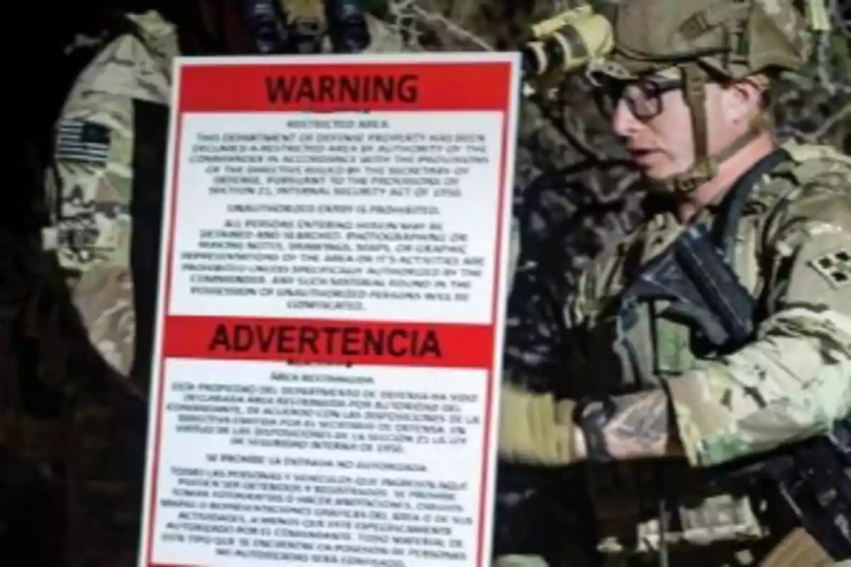 Un soldado con uniforme militar sostiene un cartel de advertencia que indica que el área es restringida.