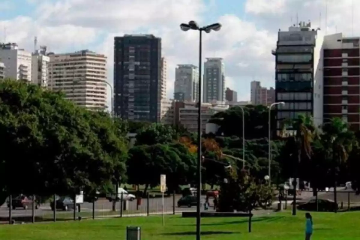 Una vista de un parque con césped verde y árboles en primer plano y edificios altos al fondo bajo un cielo parcialmente nublado.