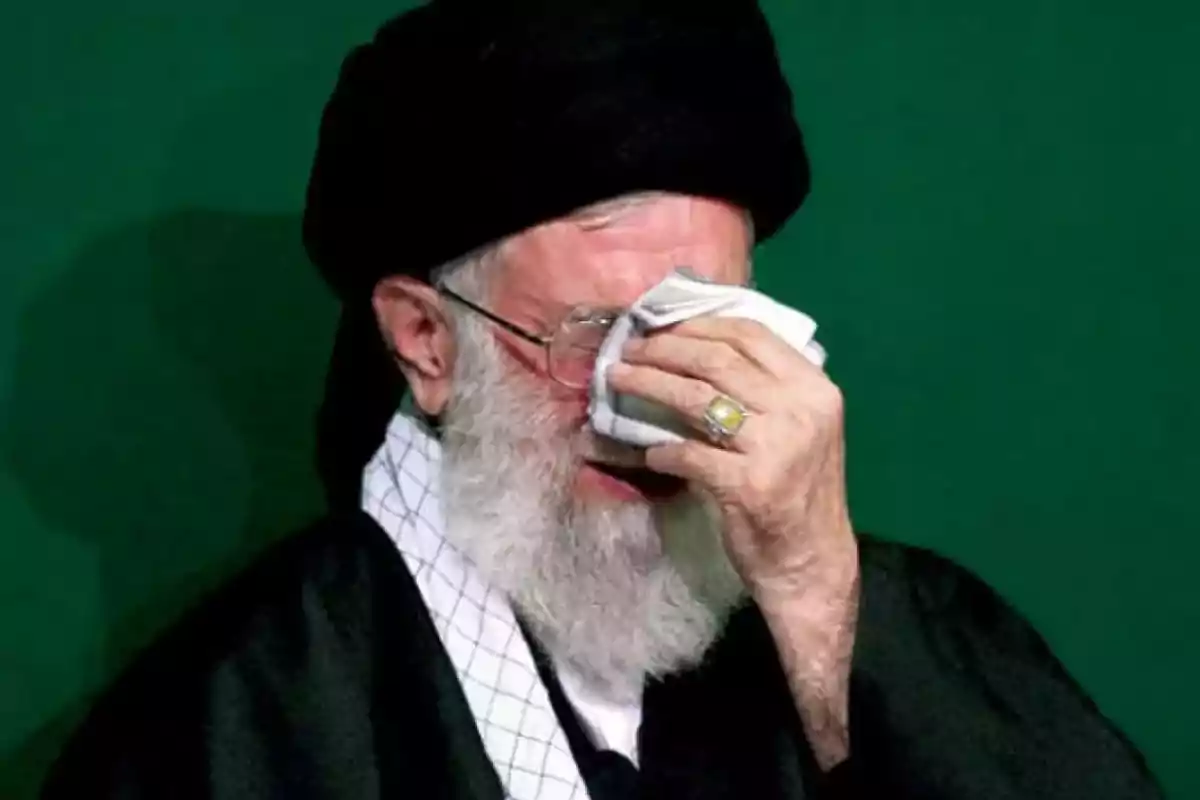 Older man with a beard and a black turban wiping his face with a white handkerchief against a green background