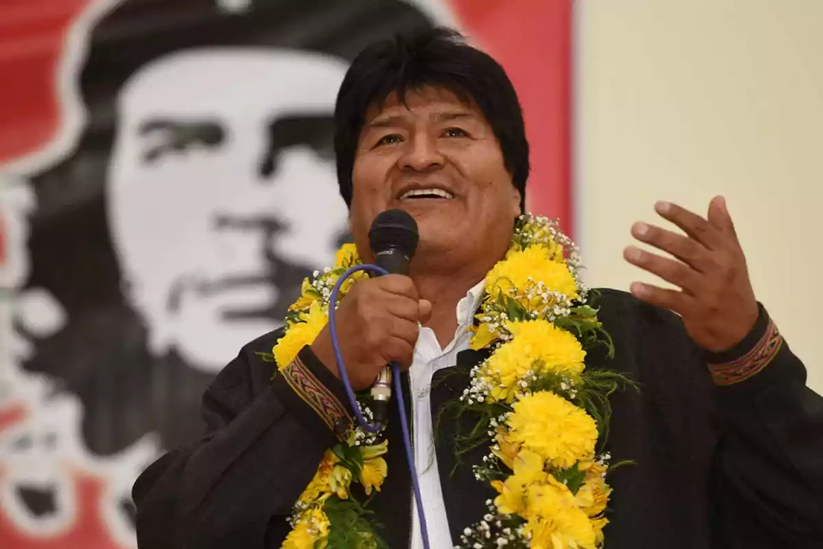 Un hombre con una guirnalda de flores amarillas habla al micrófono frente a un fondo con una imagen en blanco y negro.