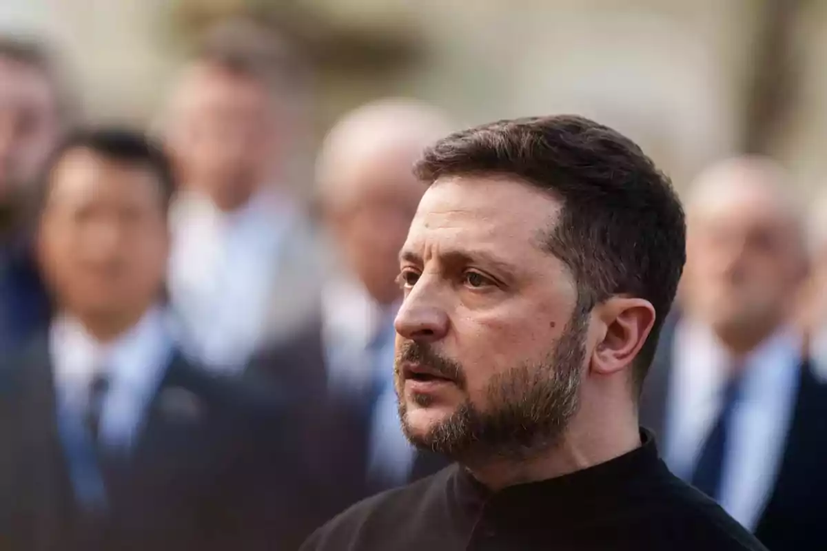 Un hombre con barba y cabello corto está de perfil en un entorno con varias personas desenfocadas al fondo. Un hombre con barba y cabello corto está de perfil en un entorno con varias personas desenfocadas al fondo.