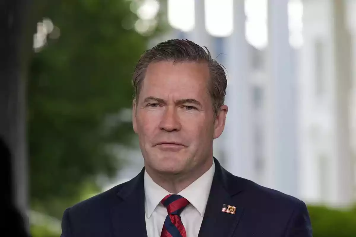 A man in a dark suit and a red and black striped tie, with a pin on the lapel, posing outdoors with a blurred background of greenery and white columns. A man in a dark suit and a red and black striped tie, with a pin on the lapel, posing outdoors with a blurred background of greenery and white columns.