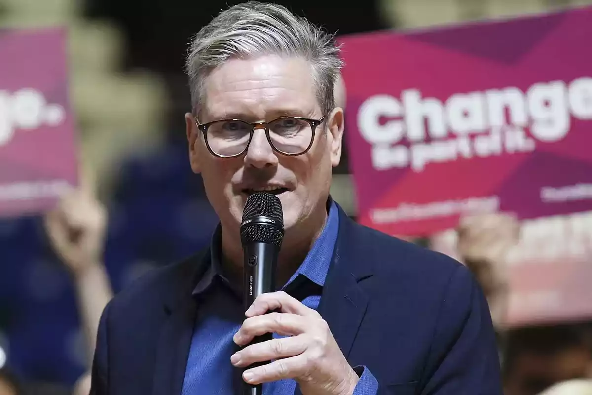 Gray-haired man with glasses speaking into a microphone at an event with signs in the background that say Change Gray-haired man with glasses speaking into a microphone at an event with signs in the background that say Change
