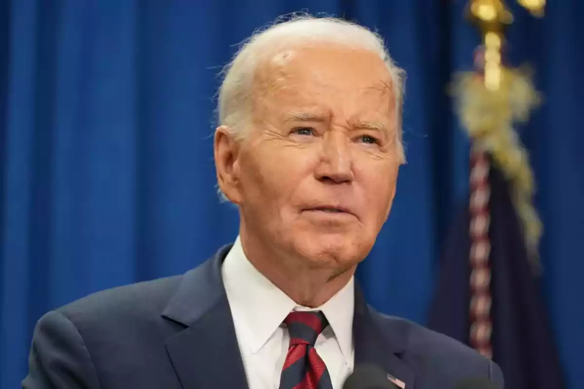 Older man with gray hair wearing a suit and striped tie in front of a blue background and a blurred flag Older man with gray hair wearing a suit and striped tie in front of a blue background and a blurred flag